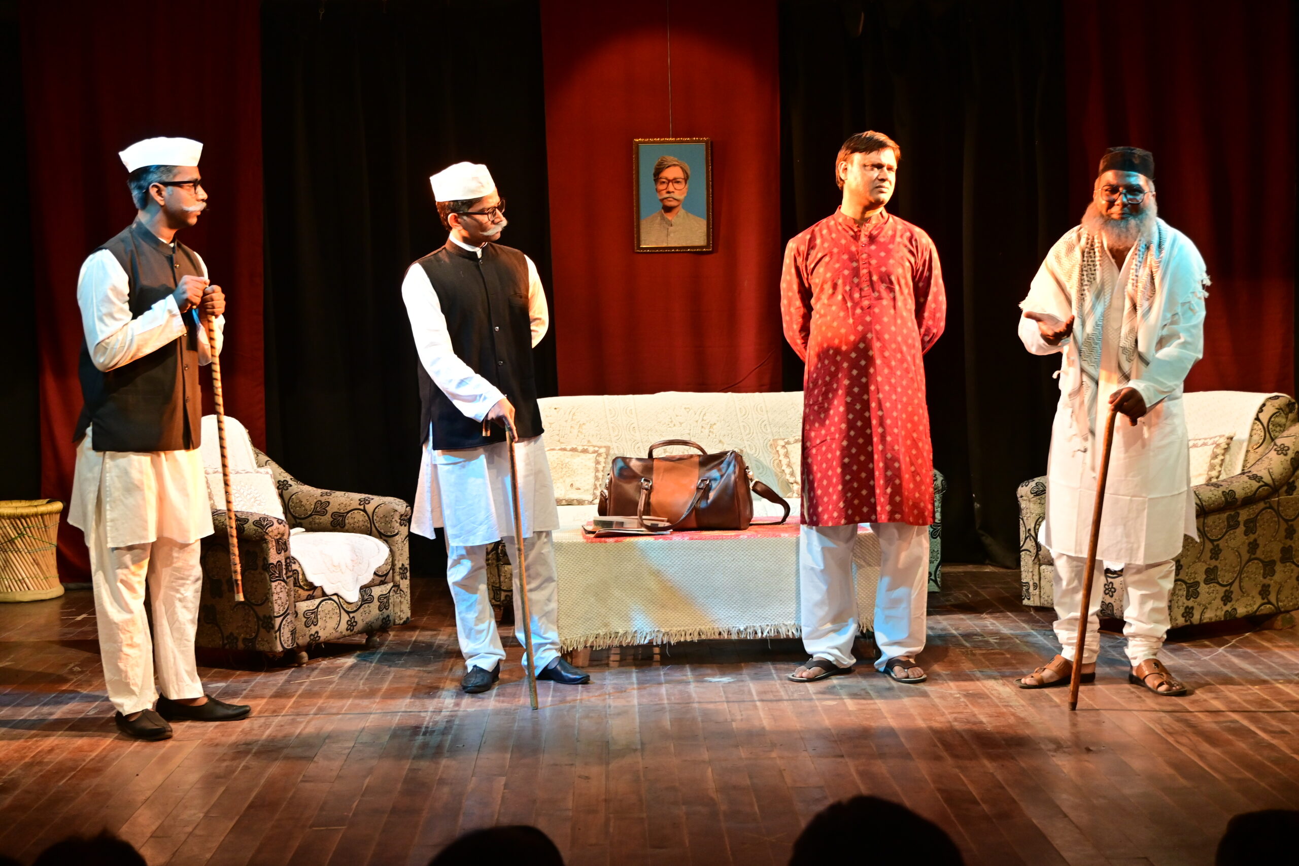 Four actors from Yayaver Rangmandal on stage in a colorful Hindi theatre performance in Lucknow, dressed in traditional Indian attire with kurtas, vests, and caps, set against a domestic scene with sofa and portrait